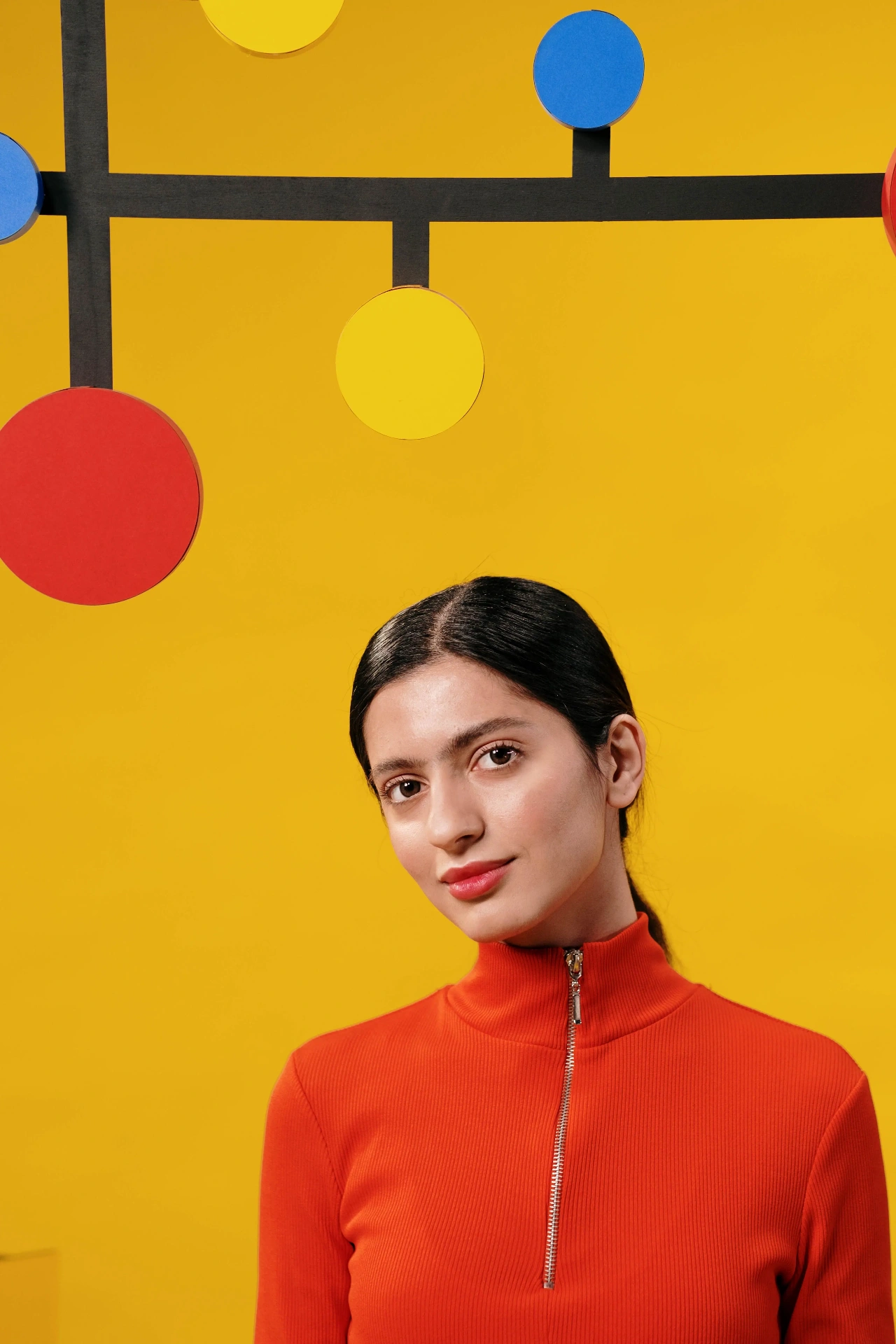Model in a red turtleneck below a yellow wall of hanging circles, by cottonbro studio.