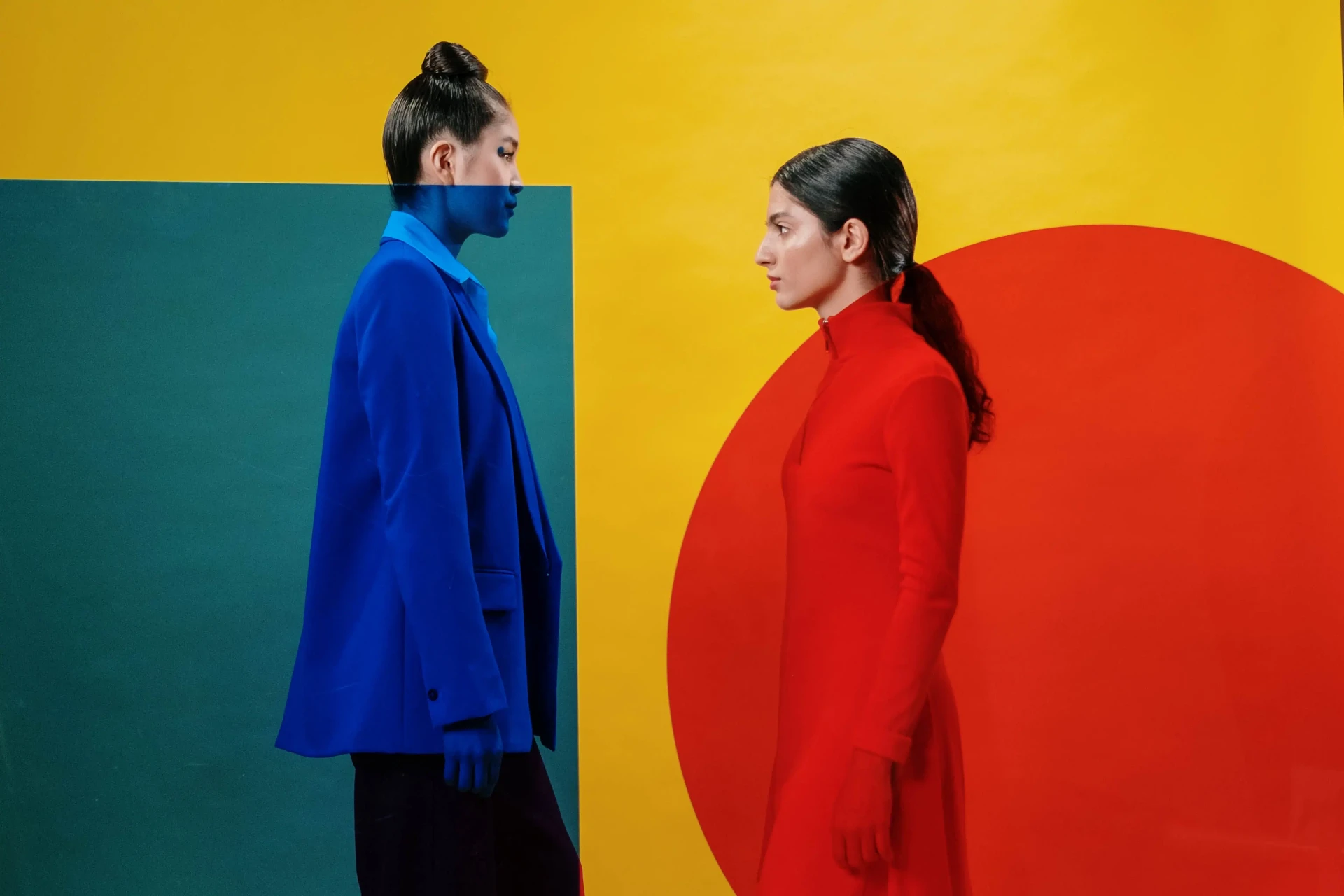 Two models in profile — one in a bright blue blazer facing another in red — against yellow and teal panels, by cottonbro studio.